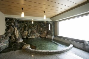 Un jacuzzi con paredes de piedra, una fuente de agua cristalina, vigas de madera en el techo y una ventana en el lado derecho.