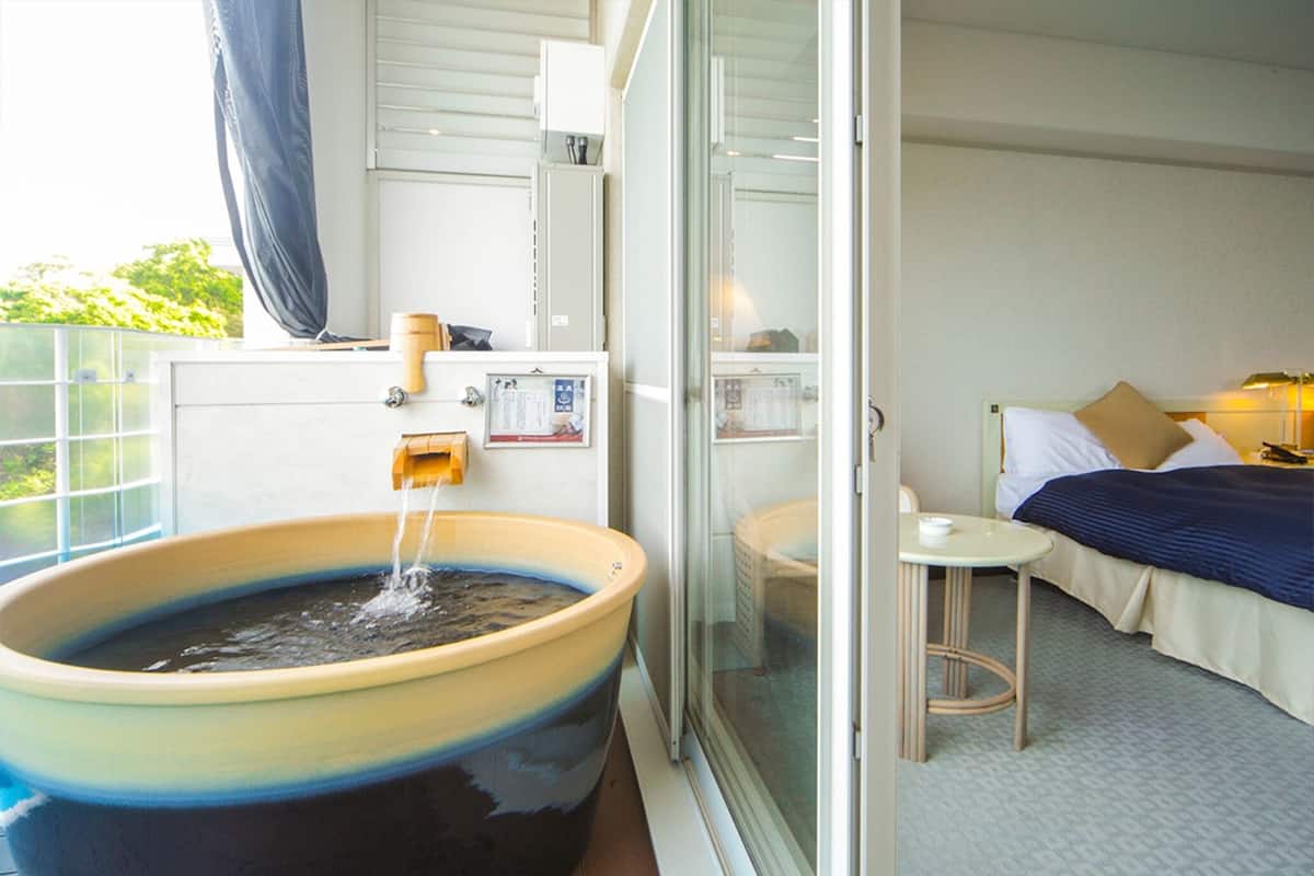 A room with a large bathtub, a bed with a blue bedspread, and a small table with a lamp.