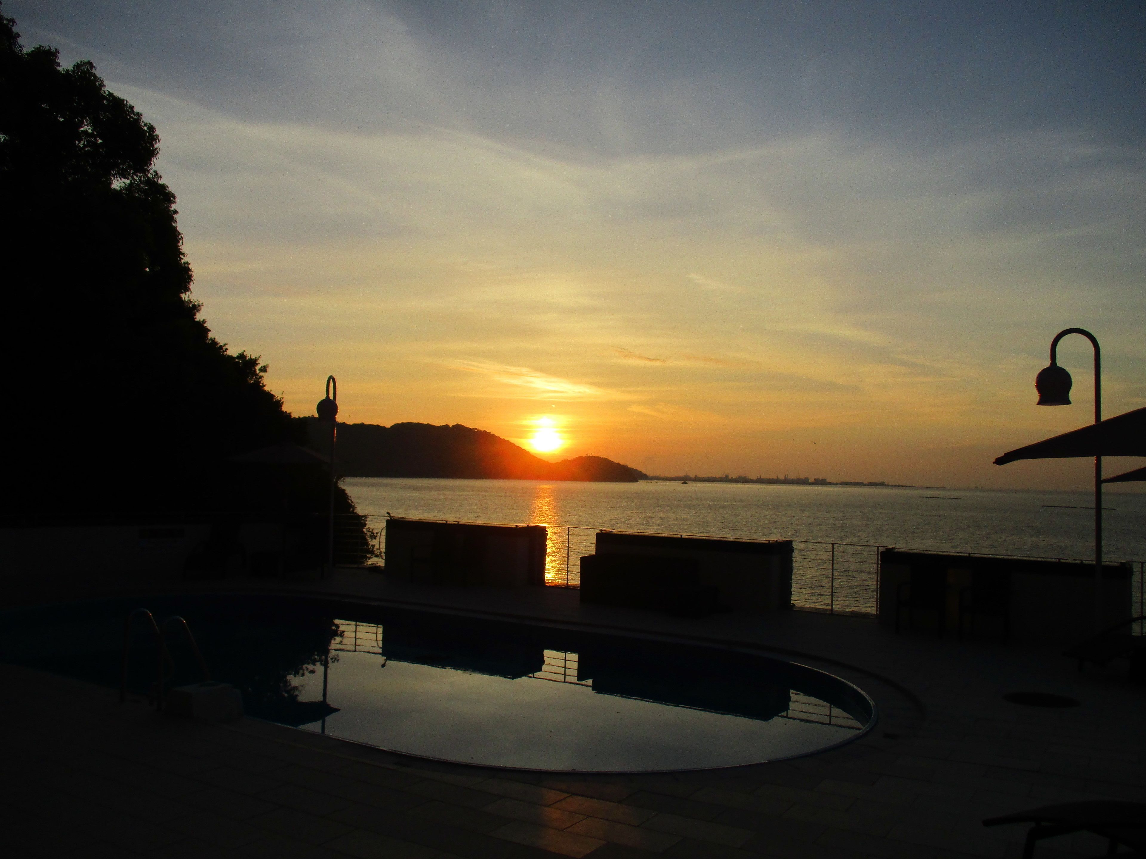 Vista de la piscina con un atardecer sobre el agua, una campana y un farol.
