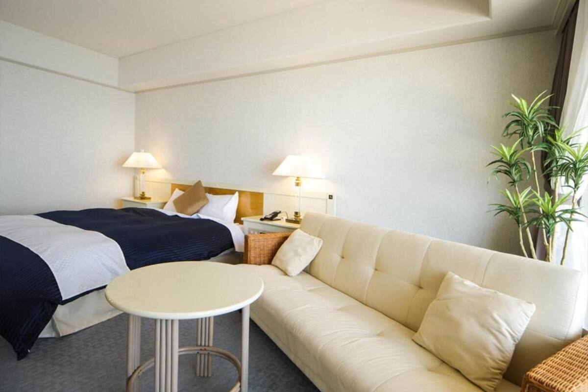 A hotel room with a bed, a sofa, a small table, two bedside lamps, a nightstand, and a potted plant.