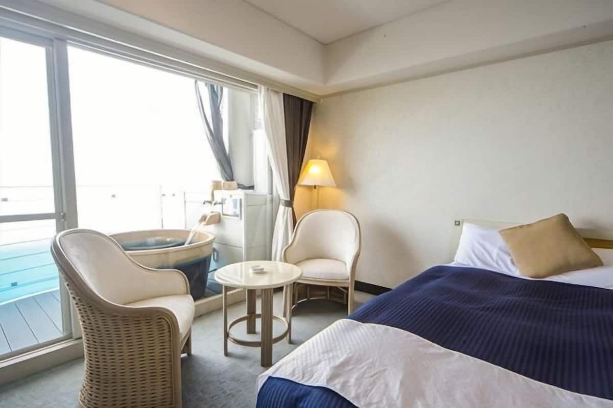 A hotel room with a bed, two chairs, a small table, a hot tub, and a view of the outside.