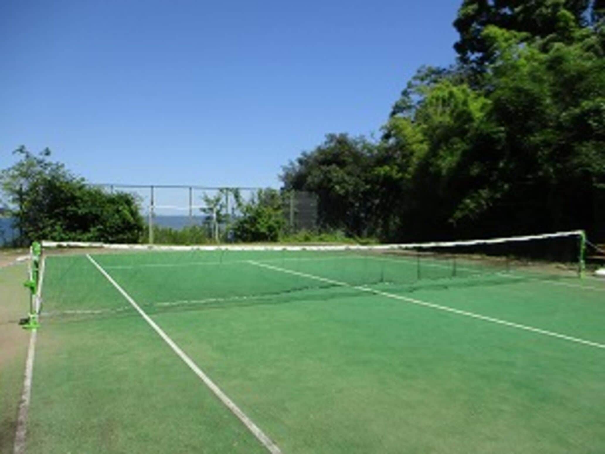 Un campo da tennis con rete, circondato da alberi e cielo sereno.