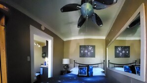 A bedroom with a bed featuring a galaxy-themed bedspread, a ceiling fan, a nightstand with a lamp, and a wall-mounted TV. - Cottonwood Hotel (Cottonwood)