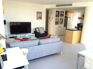 A modern living room with a grey sofa, black and white patterned cushions, a flat-screen TV, and a small coffee table. A kitchen area with a refrigerator and microwave is visible in the background. - StayCentral - Melbourne Central (Melbourne)