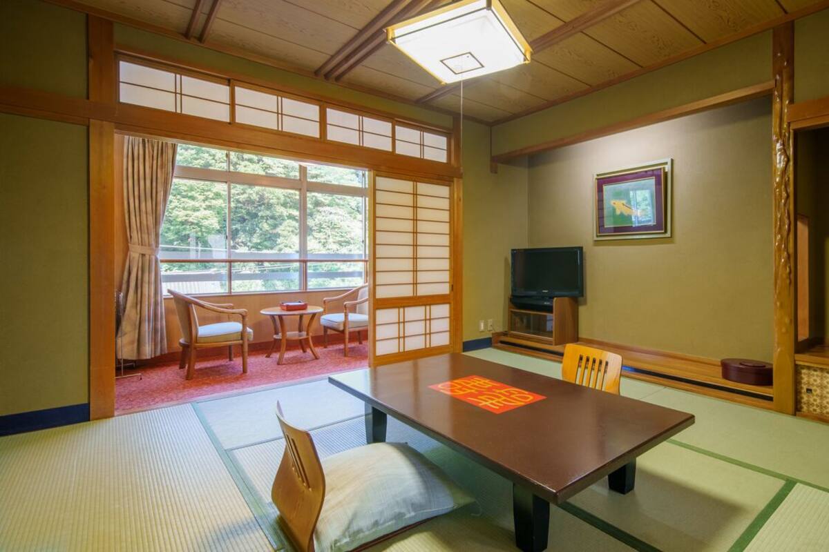 A traditional Japanese room with tatami flooring, a low table, chairs, a television, and a sliding door leading to a balcony.