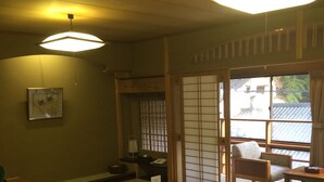 A traditional Japanese room with tatami flooring, a low table with a cushion, sliding doors, and a view of the outside. - Sento Ryokan (Toyooka)