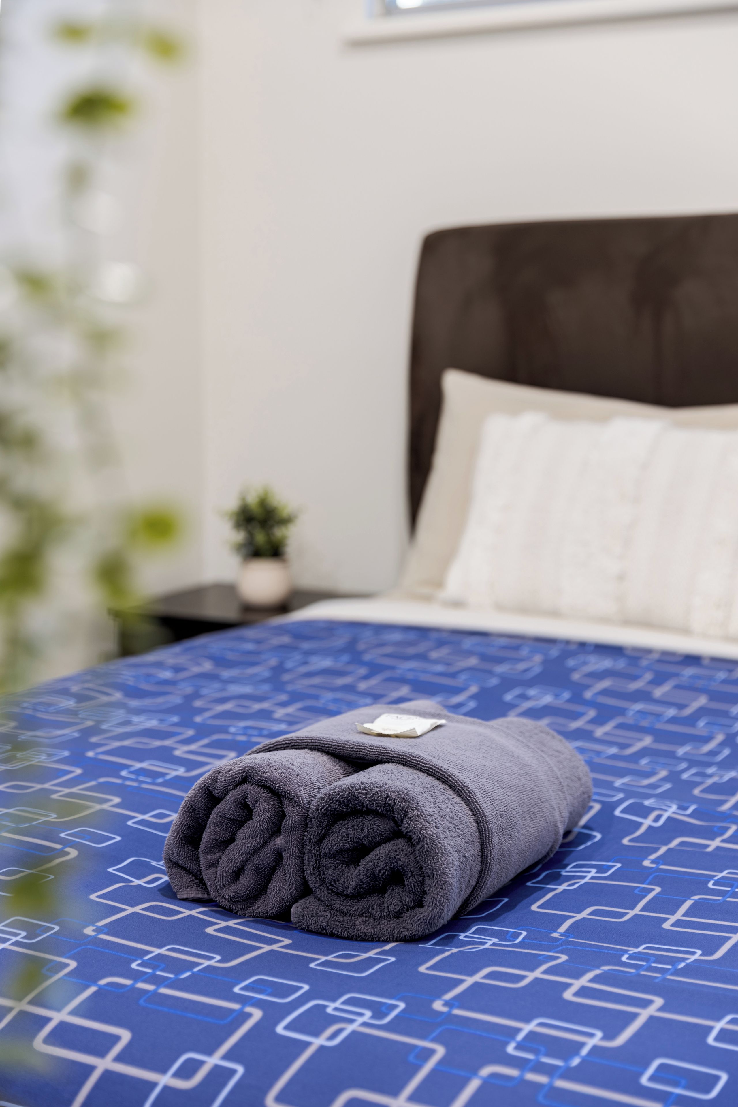A neatly made bed with a blue patterned bedspread and a rolled gray towel on top.
