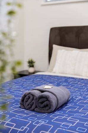 Una cama bien hecha con una colcha azul estampada y una toalla gris enrollada encima.