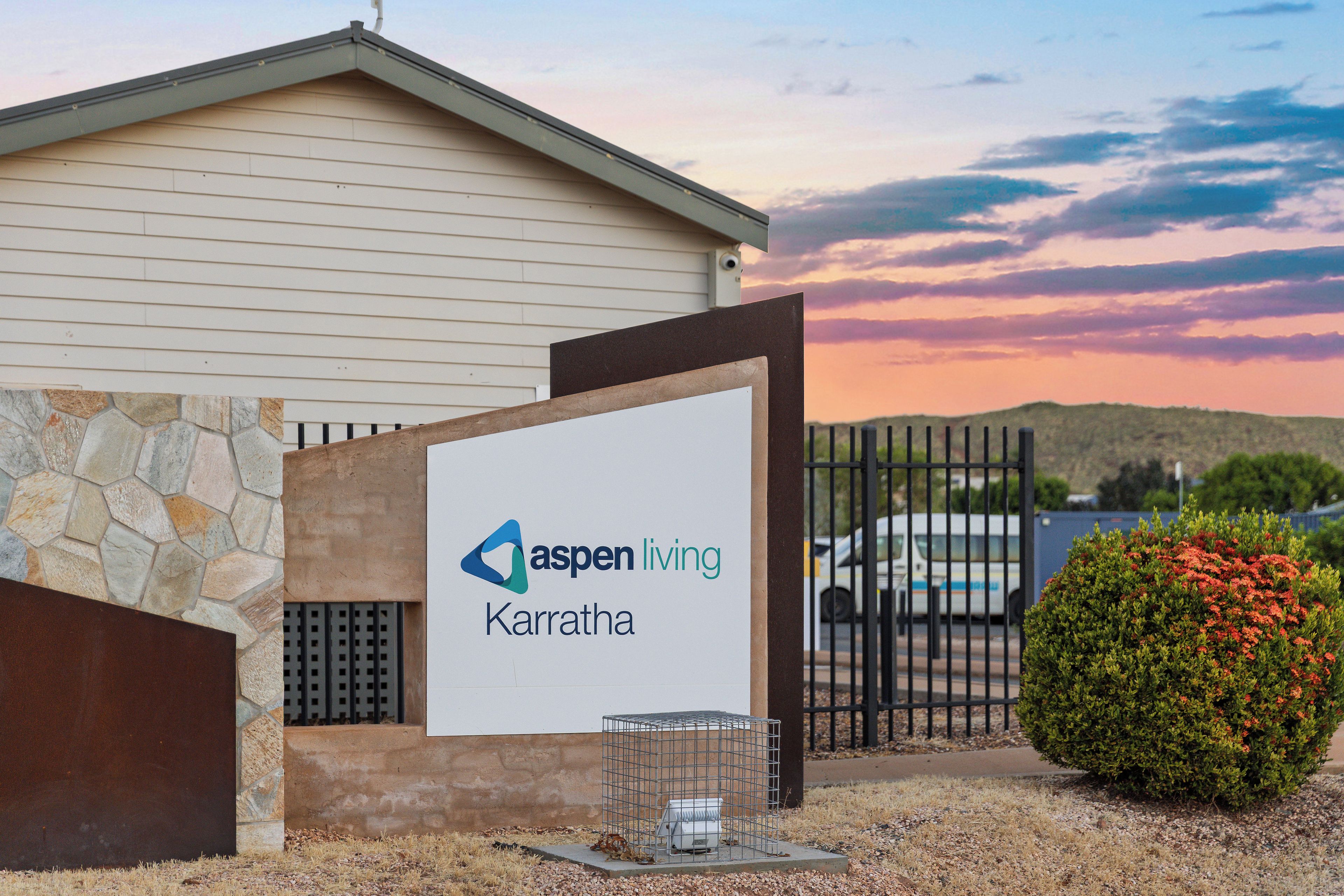 A building with a sign for Aspen Living Karratha, a gated entrance, and a small bush with red flowers.