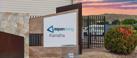 Un edificio con un letrero que dice Aspen Living Karratha, entrada con acceso restringido y un arbusto pequeño con flores rojas.