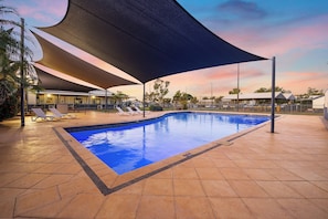 A swimming pool with a shaded area, lounge chairs, and a building in the background. - Aspen Karratha Village (Baynton)