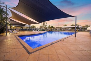 Piscina com área coberta, espreguiçadeiras e um edifício ao fundo.