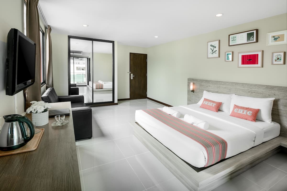A modern hotel room with a large bed, a flat-screen TV, a coffee table, and a view of the city through a glass door.