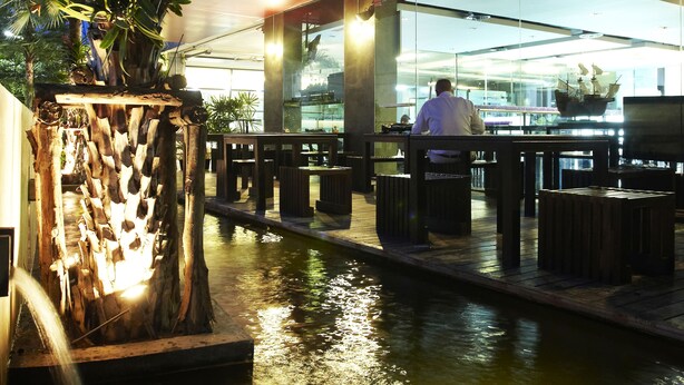 Un restaurante frente al agua con terraza al aire libre, una palmera y un edificio moderno al fondo.