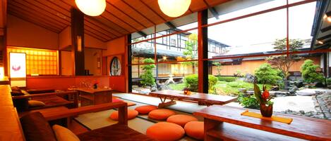 Un interior japonés tradicional con suelo de tatami, mobiliario de madera y grandes ventanales que ofrecen vistas a un jardín y edificios tradicionales.