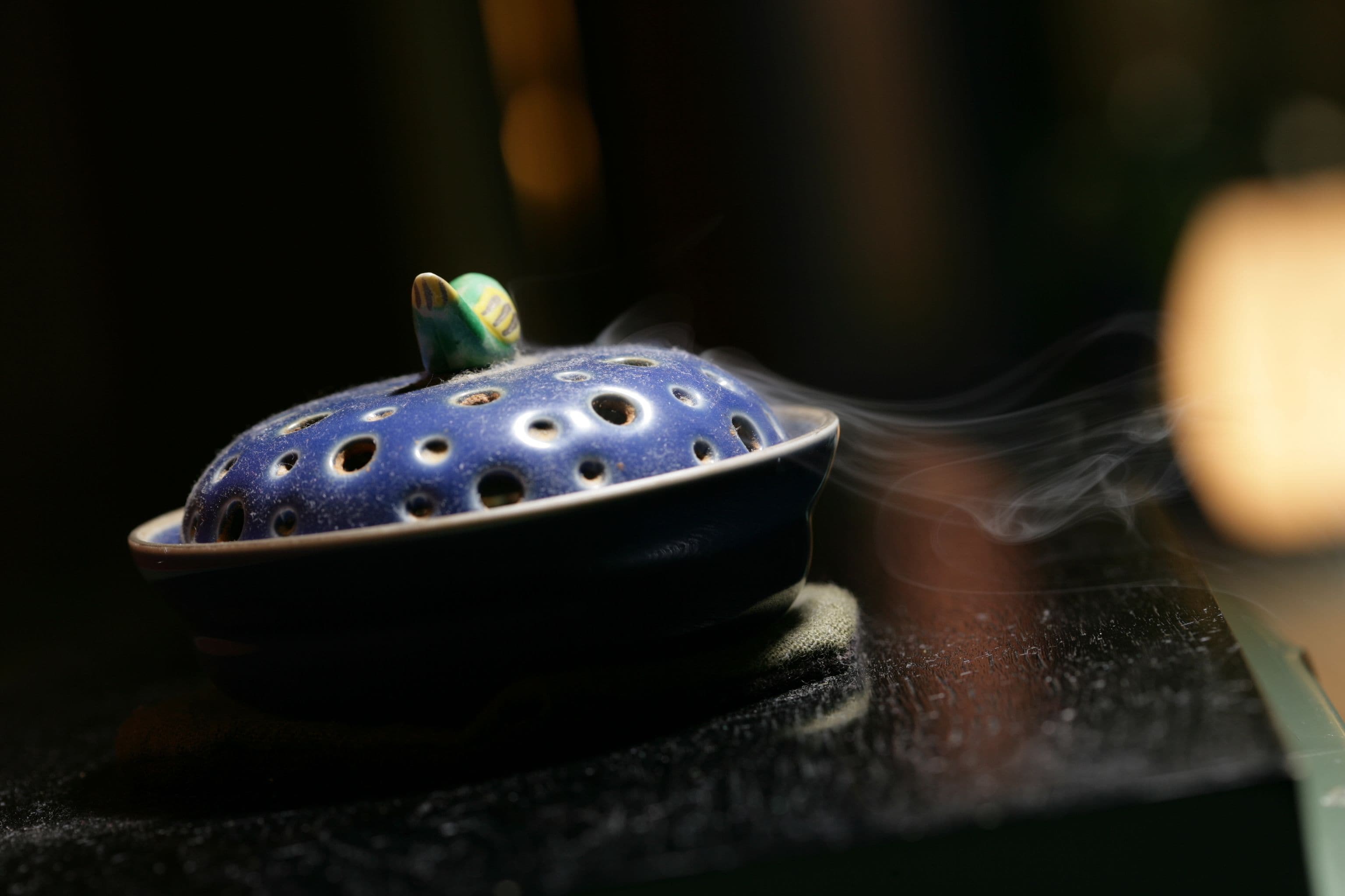 A blue incense burner with holes, emitting smoke, placed on a dark surface.