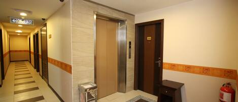 A hotel lobby with an elevator, a fire extinguisher, a trash bin, and a patterned rug.