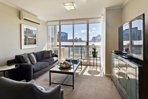 A modern living room with a sofa, coffee table, and TV, large windows with a city view, and a balcony with a table and chairs. - StayCentral - Cityside on Whiteman (Southbank)