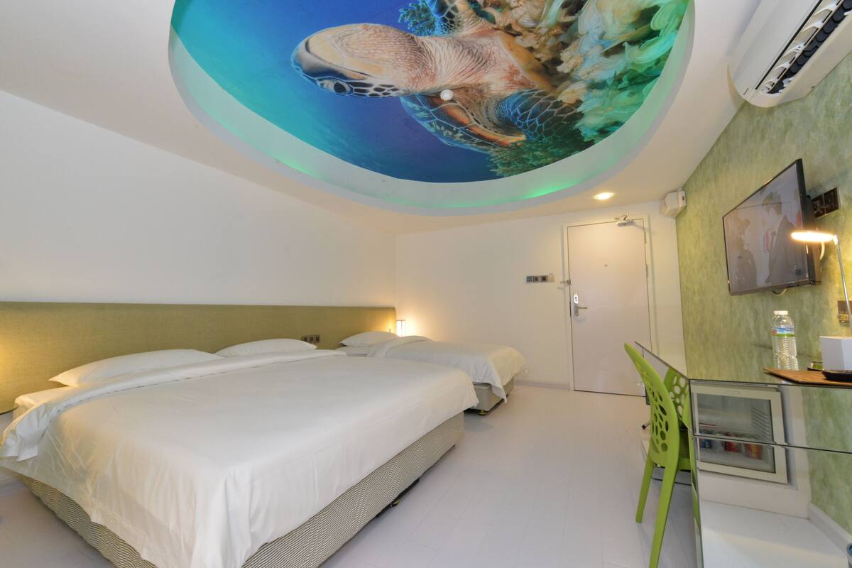 Habitación de hotel con una cama grande, una silla verde, un escritorio con una computadora y un mural en el techo con la imagen de una tortuga.