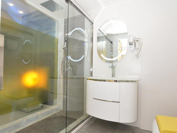 Un baño moderno con ducha envuelta en vidrio, tocador blanco con espejo y una banca amarilla.