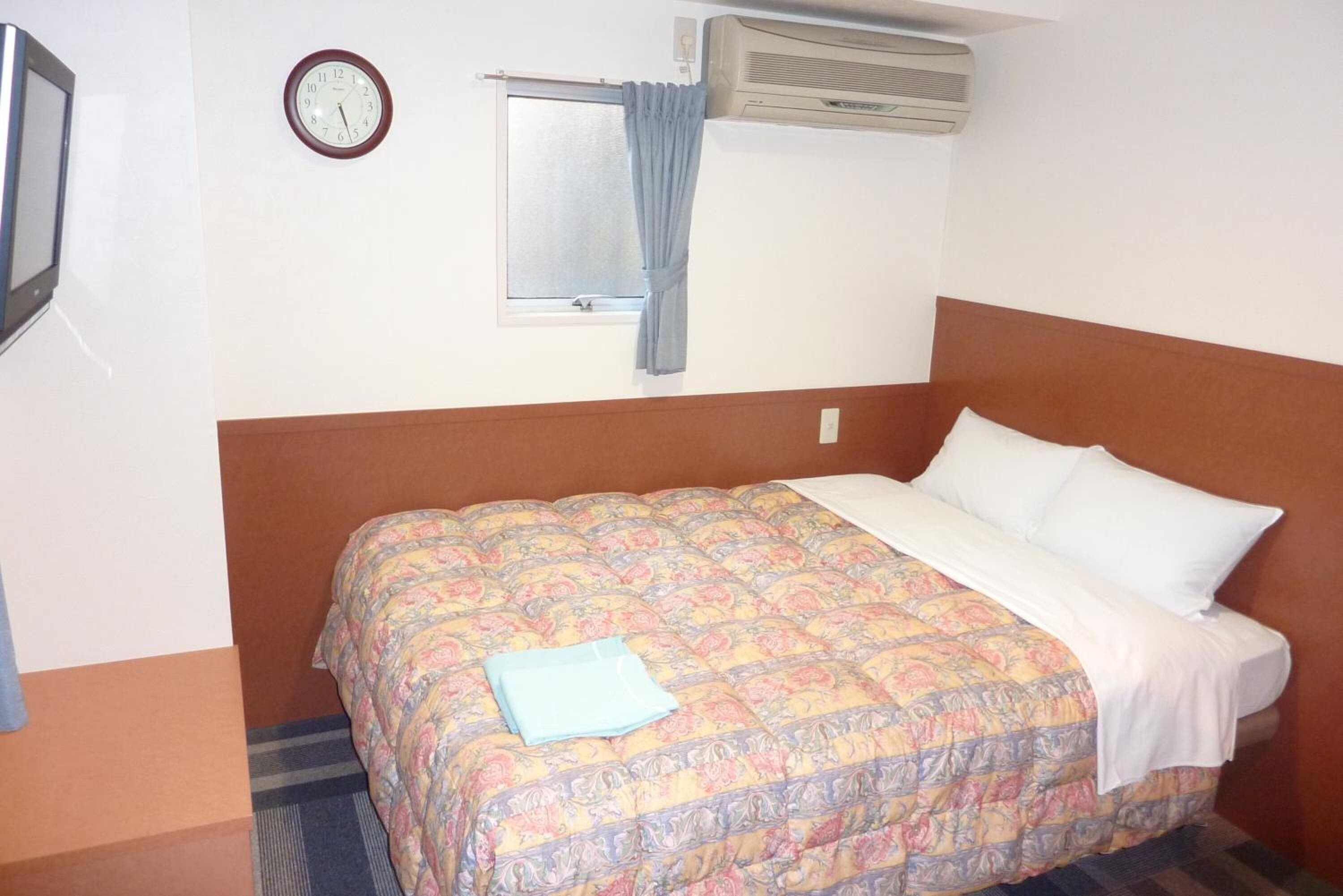 A hotel room with a bed, a clock, an air conditioner, and a window with curtains.