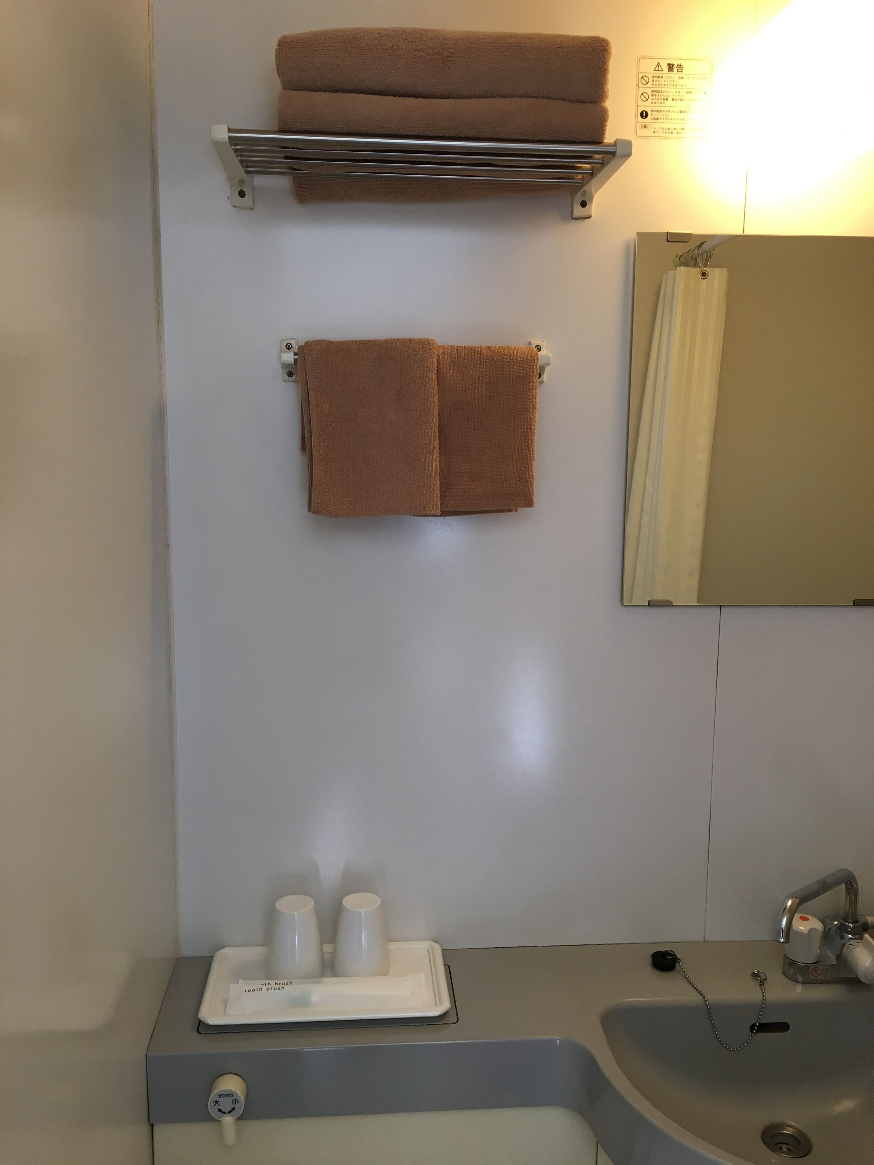 A bathroom with a towel rack holding two towels, a shelf above the sink, a mirror, and a tray with cups and a toothbrush.