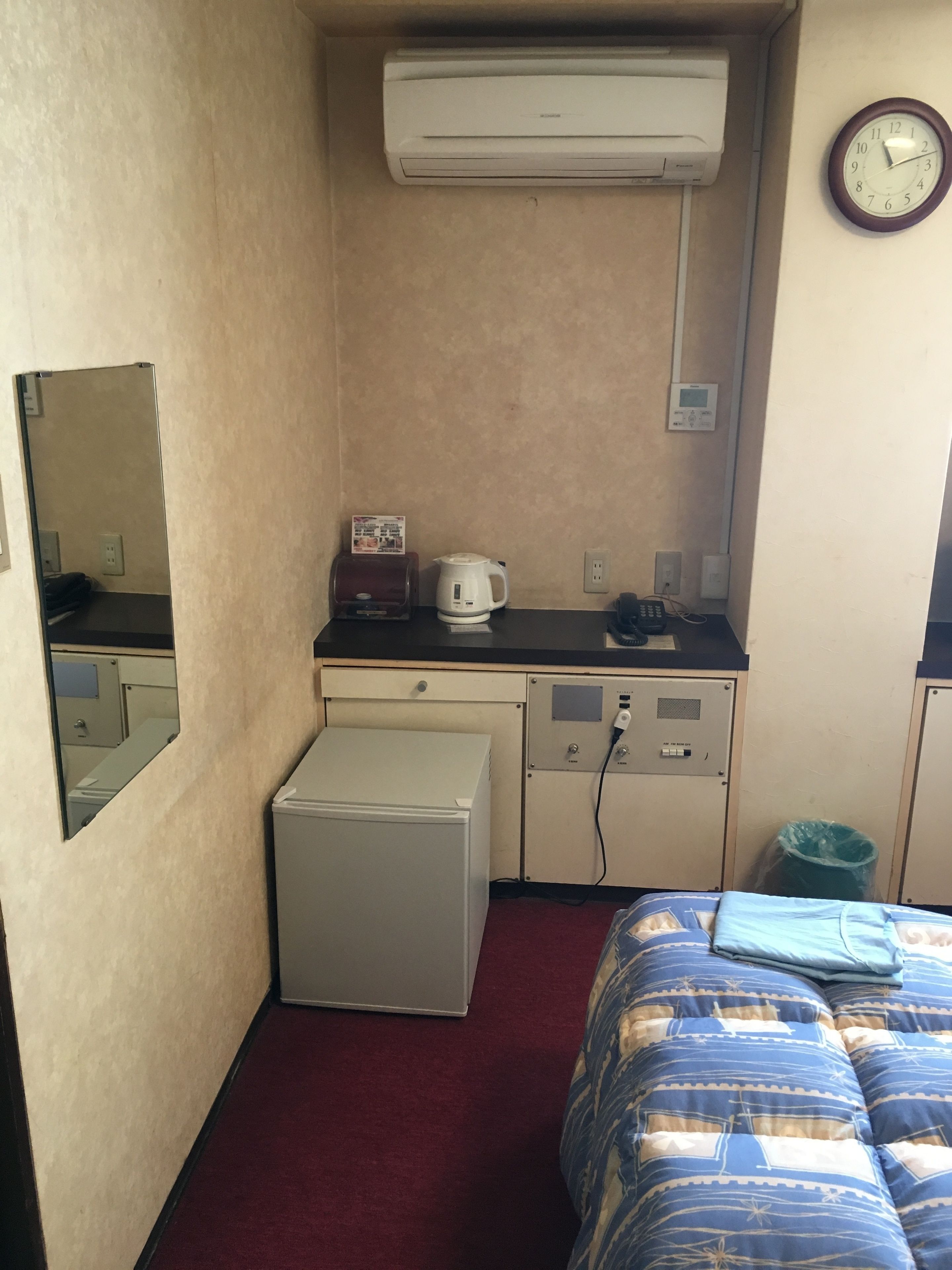 A compact hotel room with a kitchenette, a bed, a small refrigerator, a coffee maker, and a wall clock.