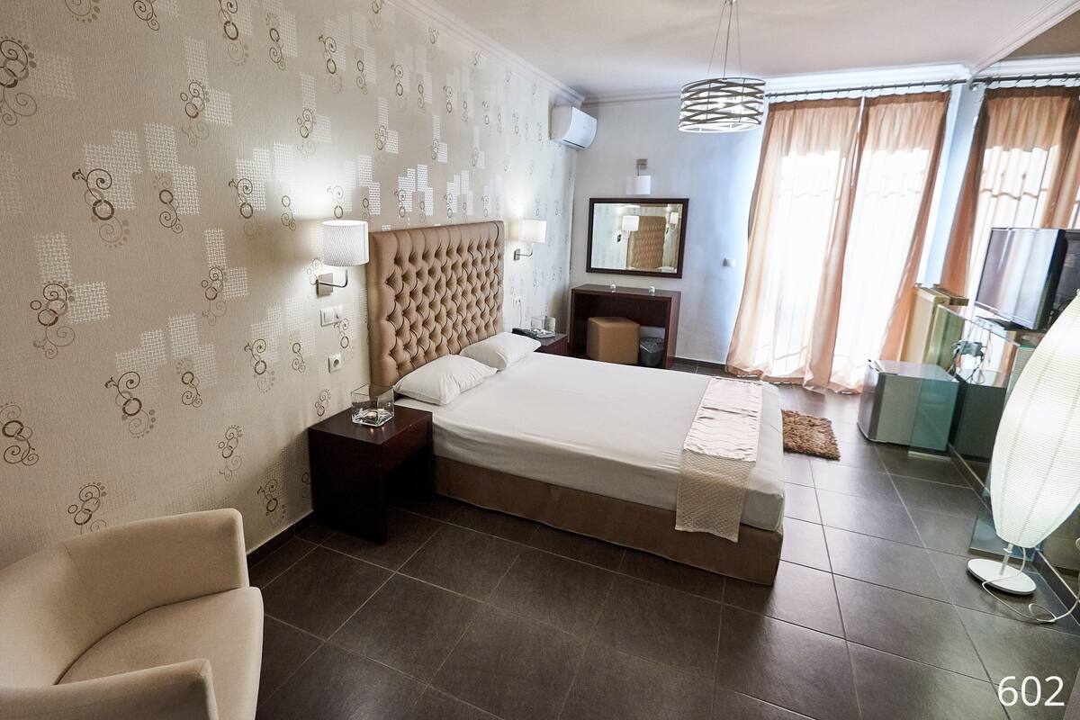 A hotel room with a large bed, a chair, a desk, a mirror, a TV, and a window with curtains.