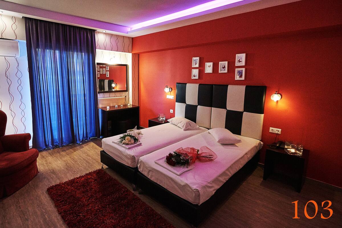 A hotel room with a checkered headboard, two beds, a red armchair, a red rug, and a large window with blue curtains.