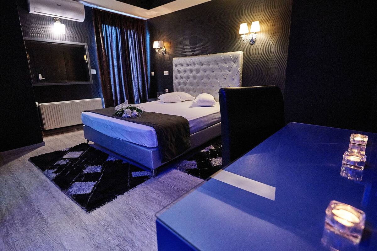 A modern hotel room with a large bed, a flat-screen TV, a bedside table with candles, and a dark-colored wall with a textured pattern.