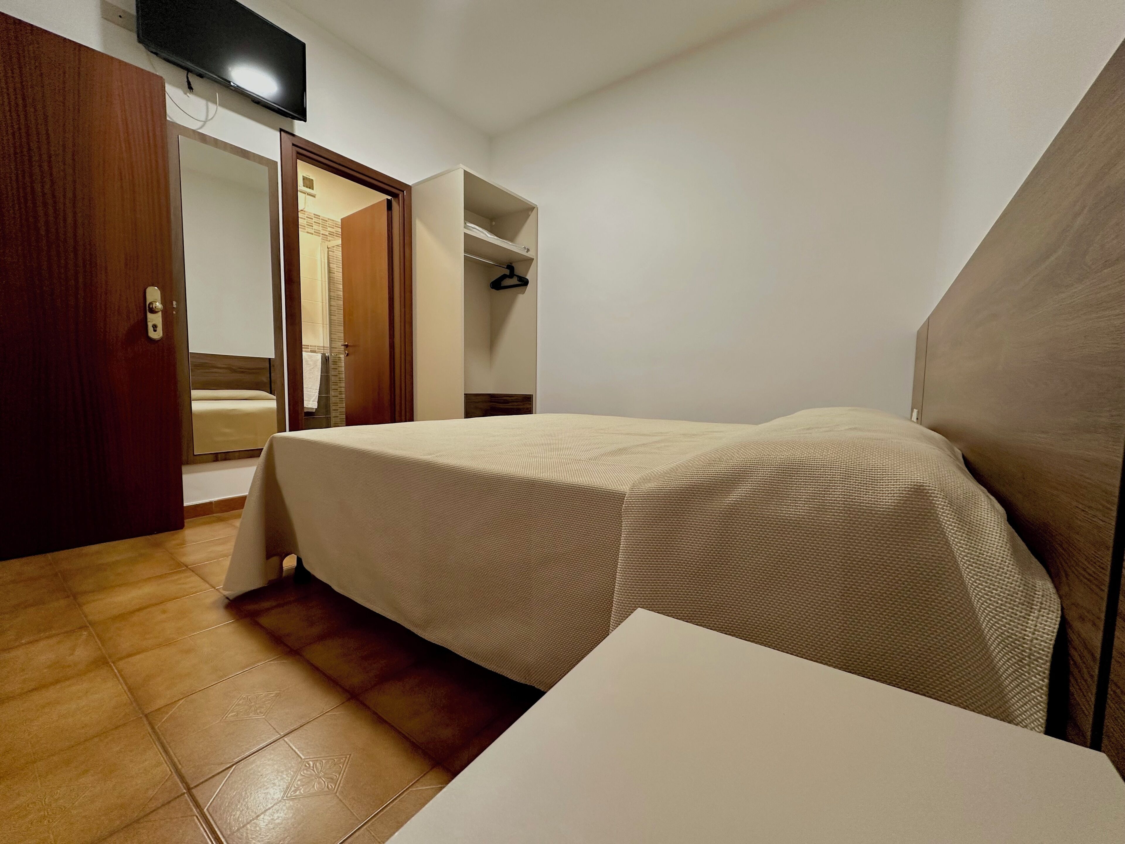 Una camera d'albergo con un letto, un armadio in legno, un televisore montato a parete e un bagno visibile attraverso una porta aperta.