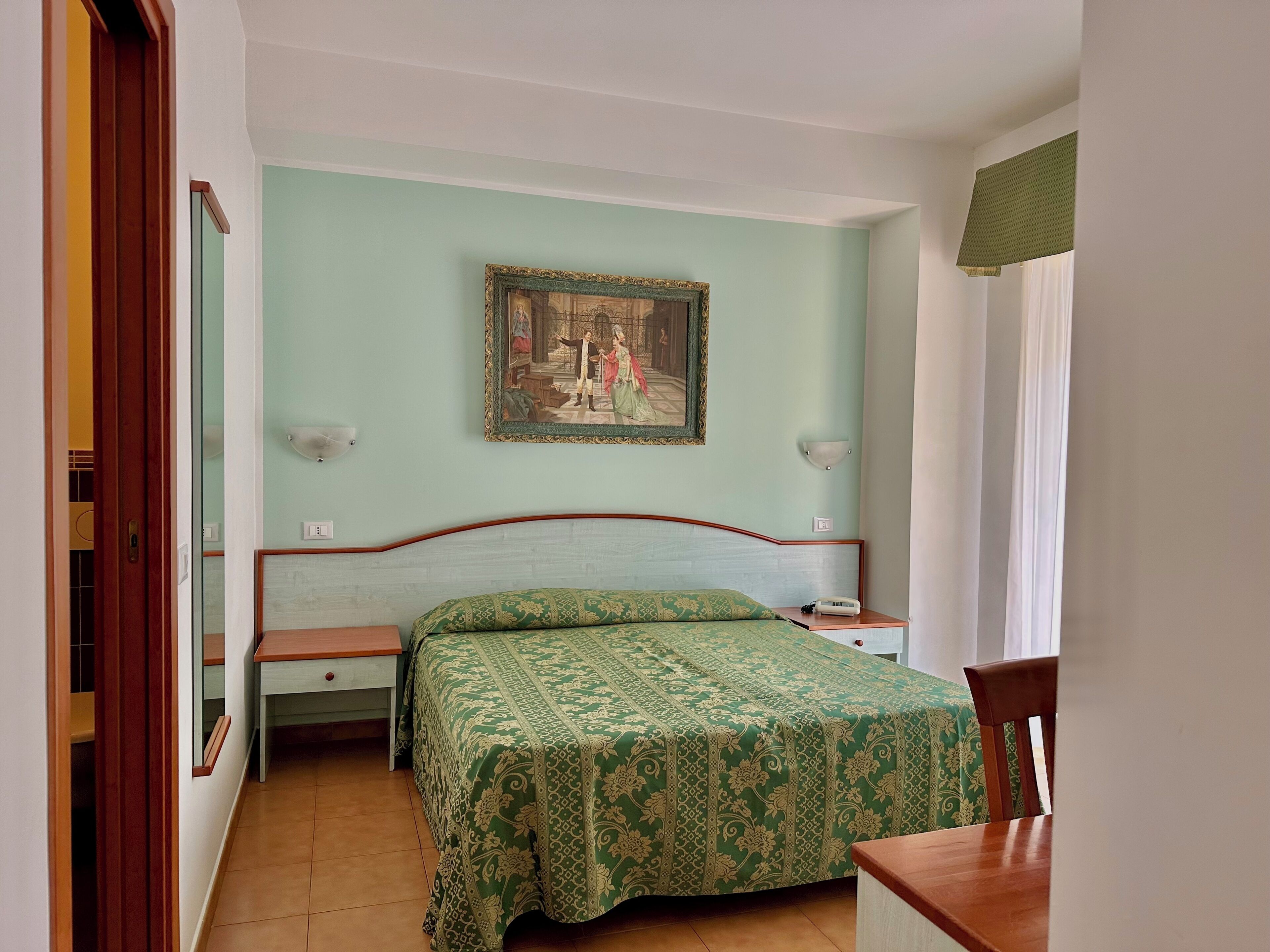 Une chambre à coucher avec un lit, des tables de chevet, un miroir, un tableau accroché au mur et une fenêtre avec des rideaux.