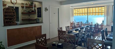 A restaurant with a bar area, multiple tables, chairs, and a view of the sea.