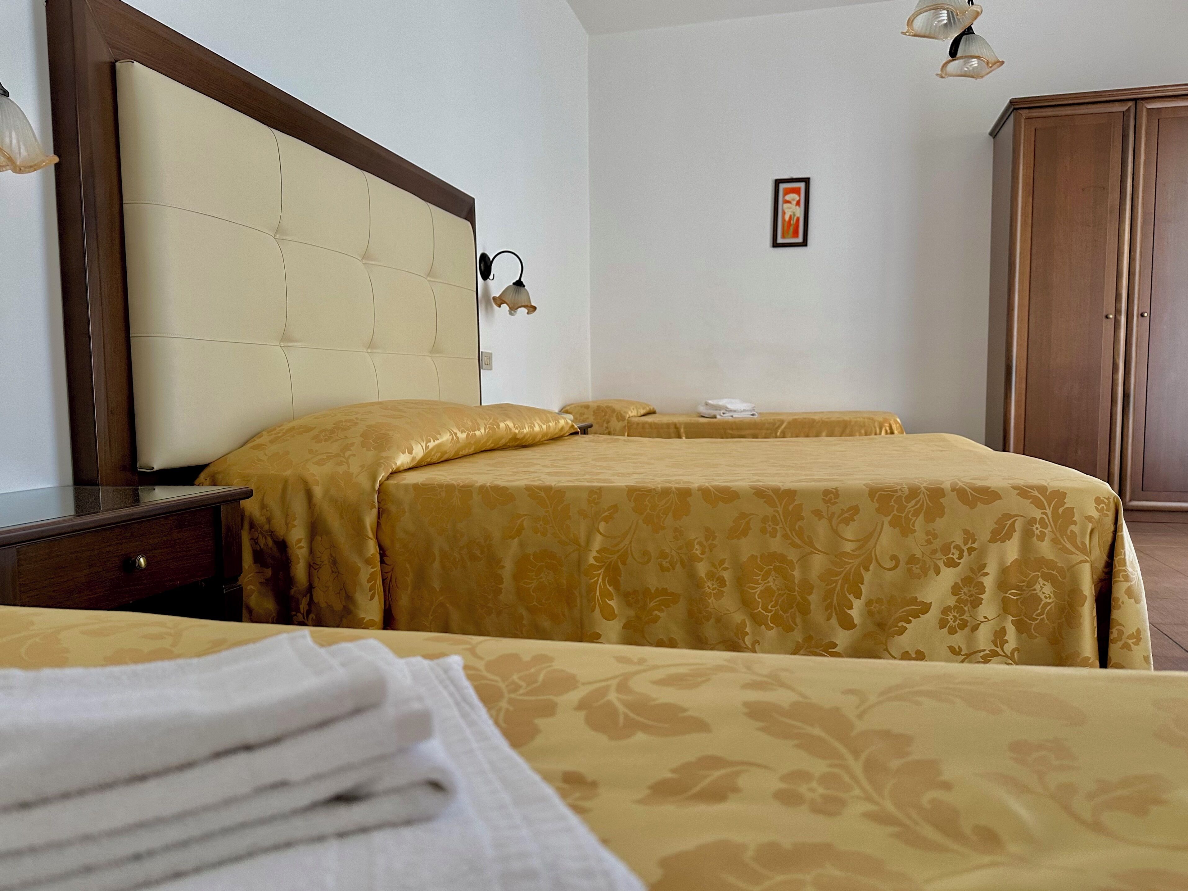 A hotel room with two beds, a wooden wardrobe, a bedside table, and a framed picture on the wall.