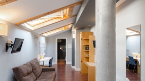 Una habitación moderna estilo loft con techo inclinado, vigas de madera, un pilar de hormigón, un sofá marrón y una zona de escritorio con una silla.