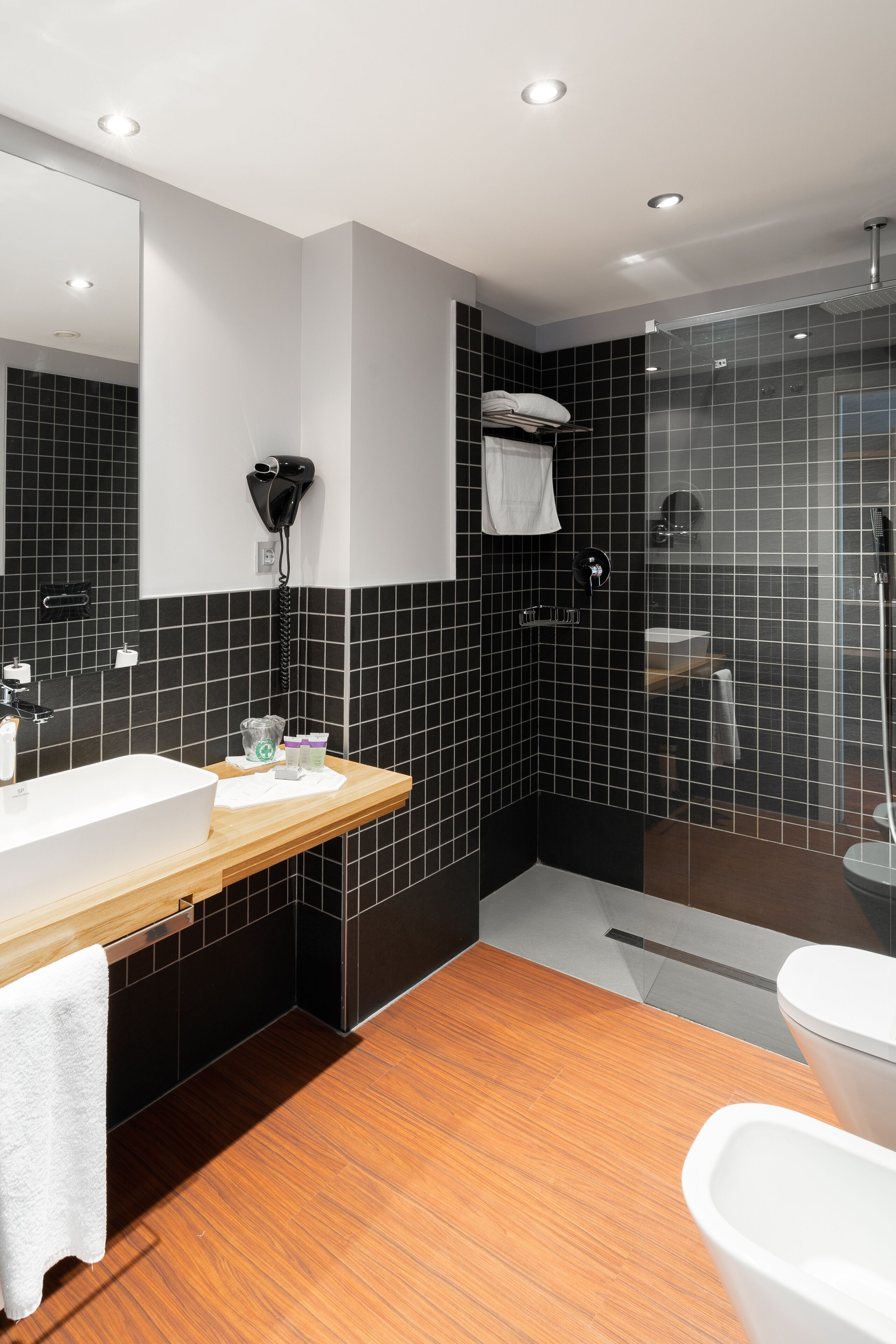 A modern bathroom with a wooden vanity, a large mirror, a glass shower enclosure, and black tiled walls.