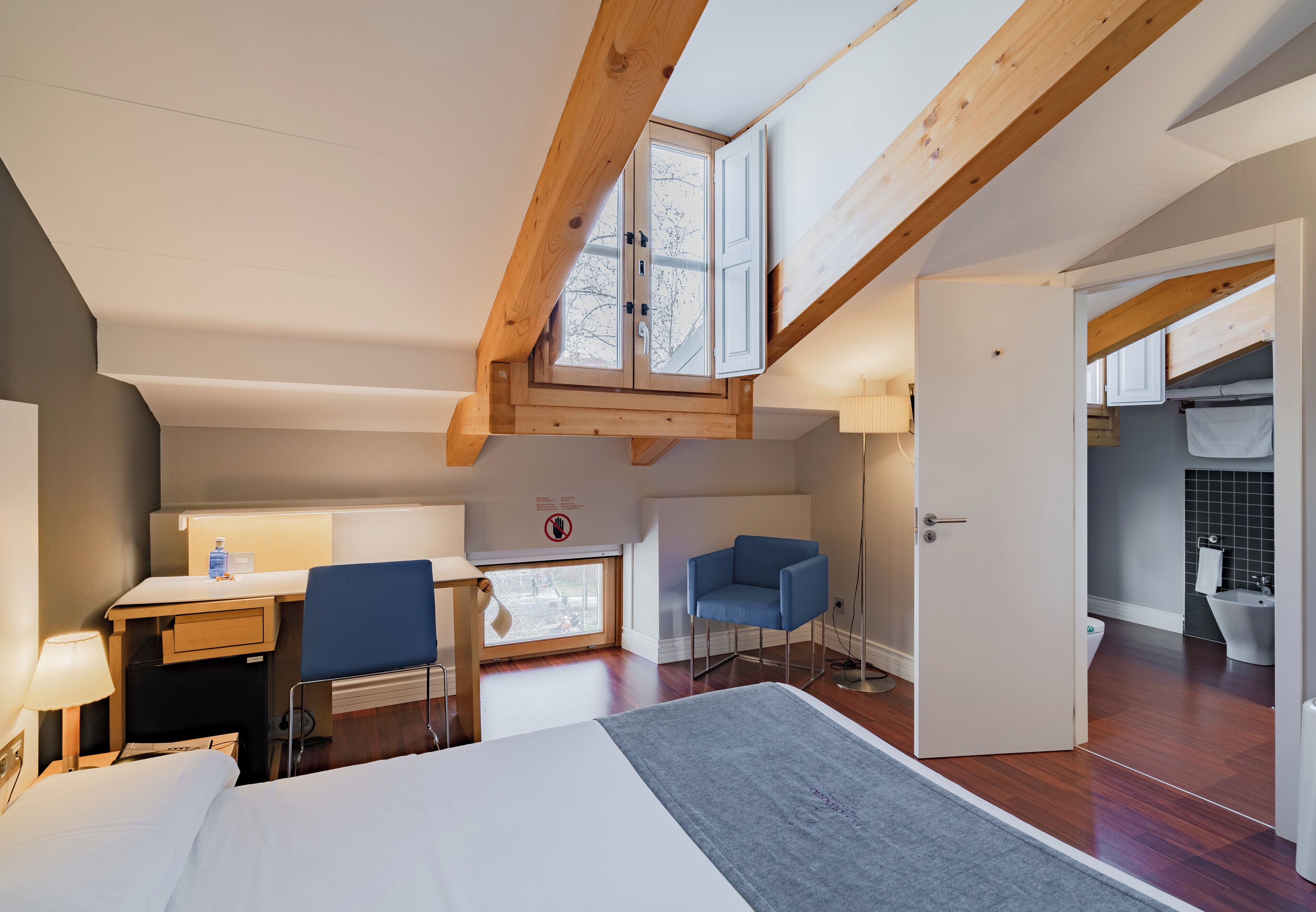 Quarto de hotel compacto com cama, escrivaninha, cadeira e uma pequena cozinha.