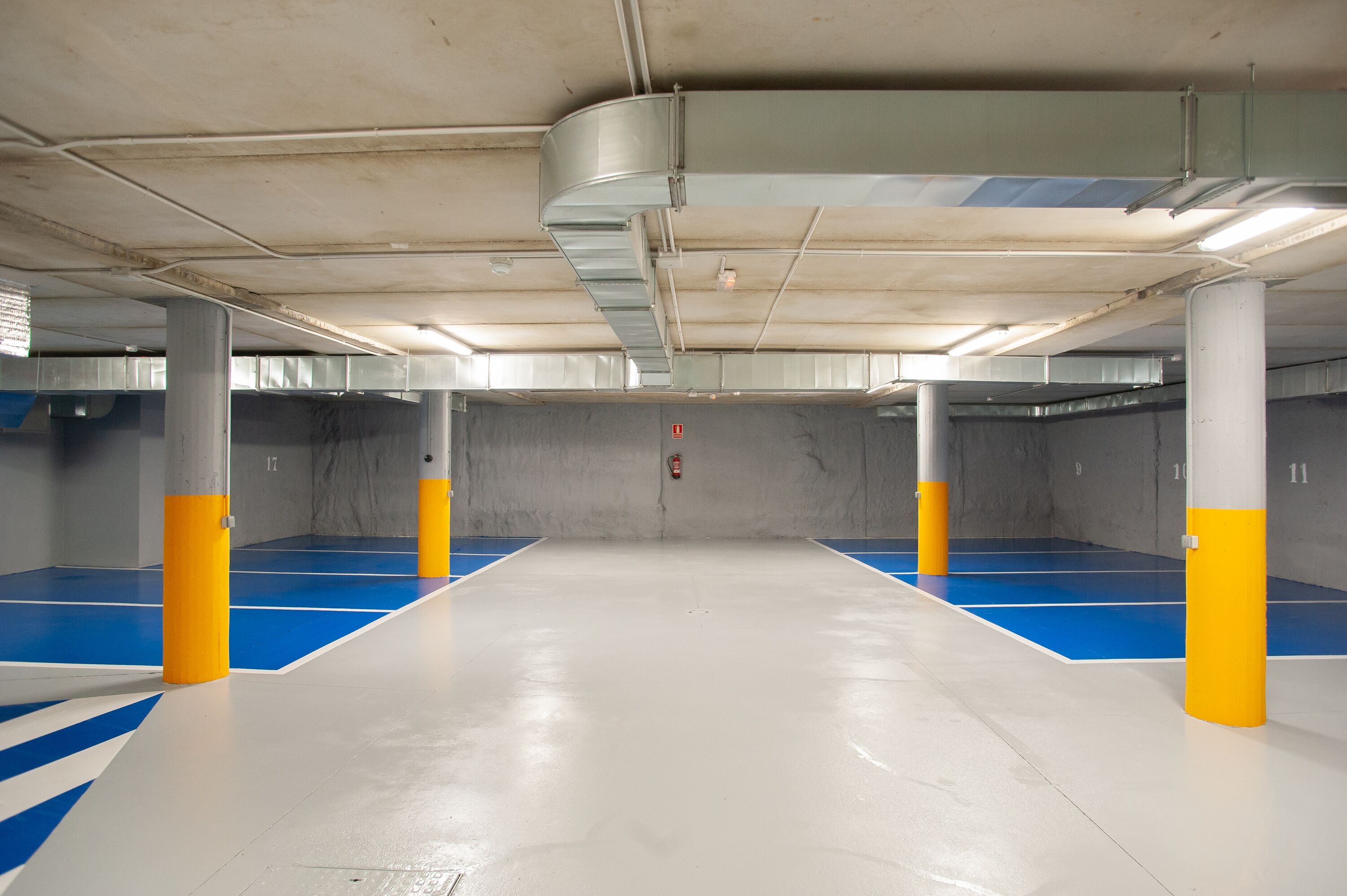 Un estacionamiento interior con pilares de hormigón, marcas en el suelo azul y blanco, e iluminación colgante.