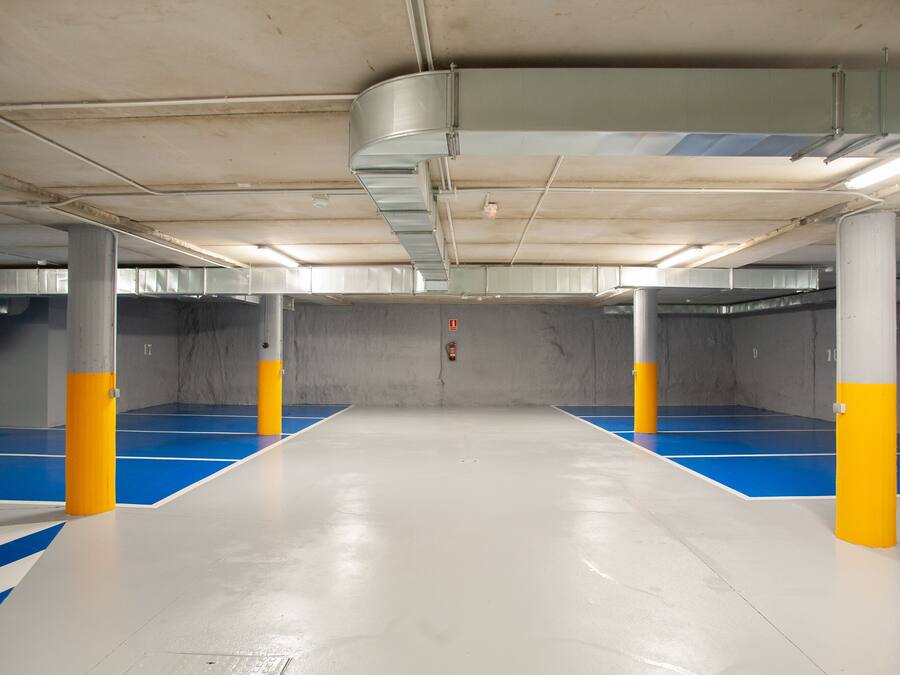 Un estacionamiento interior con pilares de hormigón, marcas en el suelo azul y blanco, e iluminación colgante.