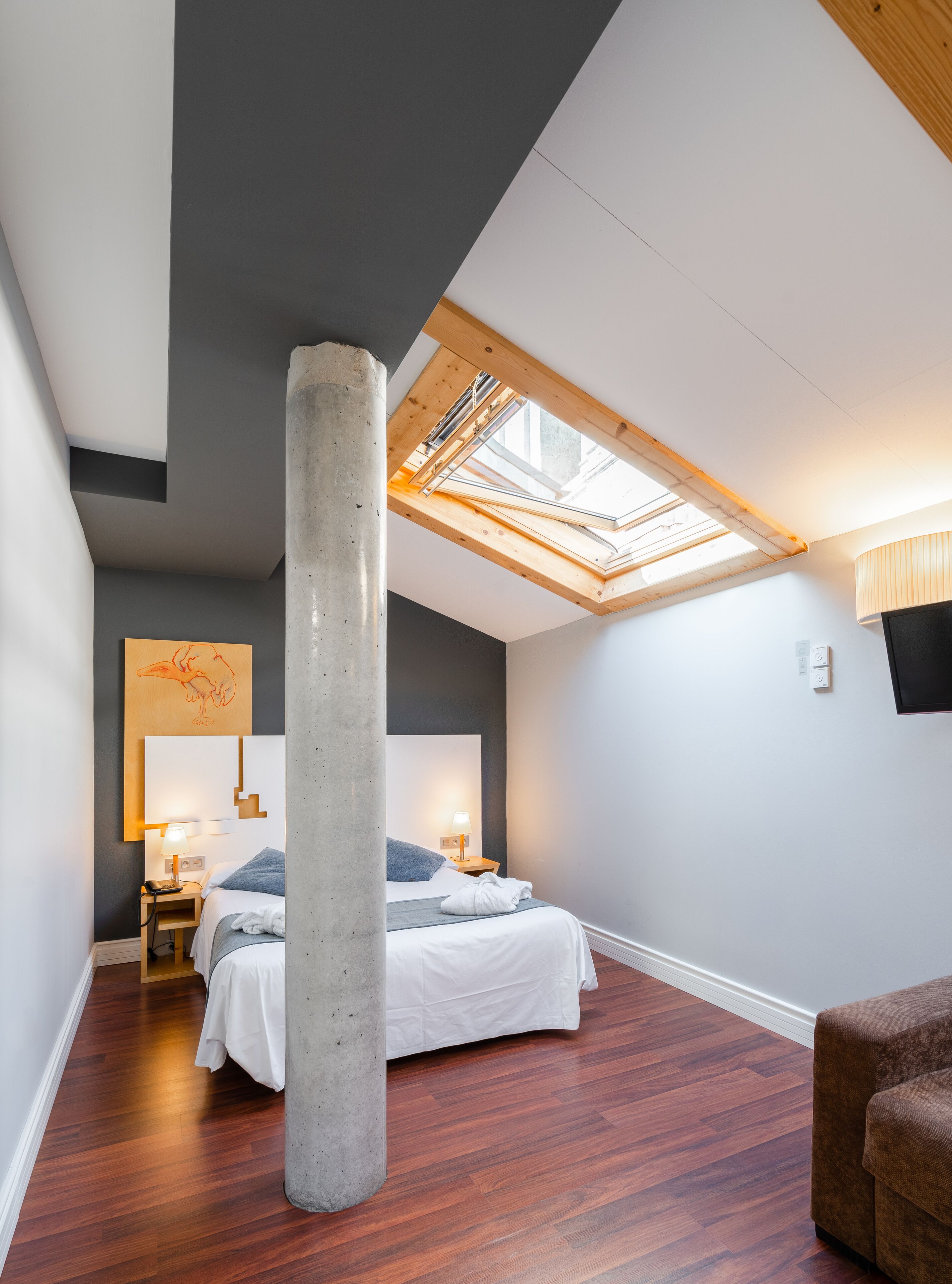 A modern bedroom with a skylight, a large concrete pillar, a bed with white linens, a wooden nightstand, and a brown sofa.