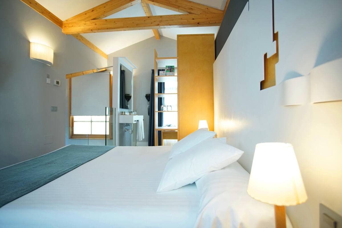A hotel room with a large bed, a bedside lamp, a wooden ceiling, a bathroom with a mirror, and a shelf with books and glasses.