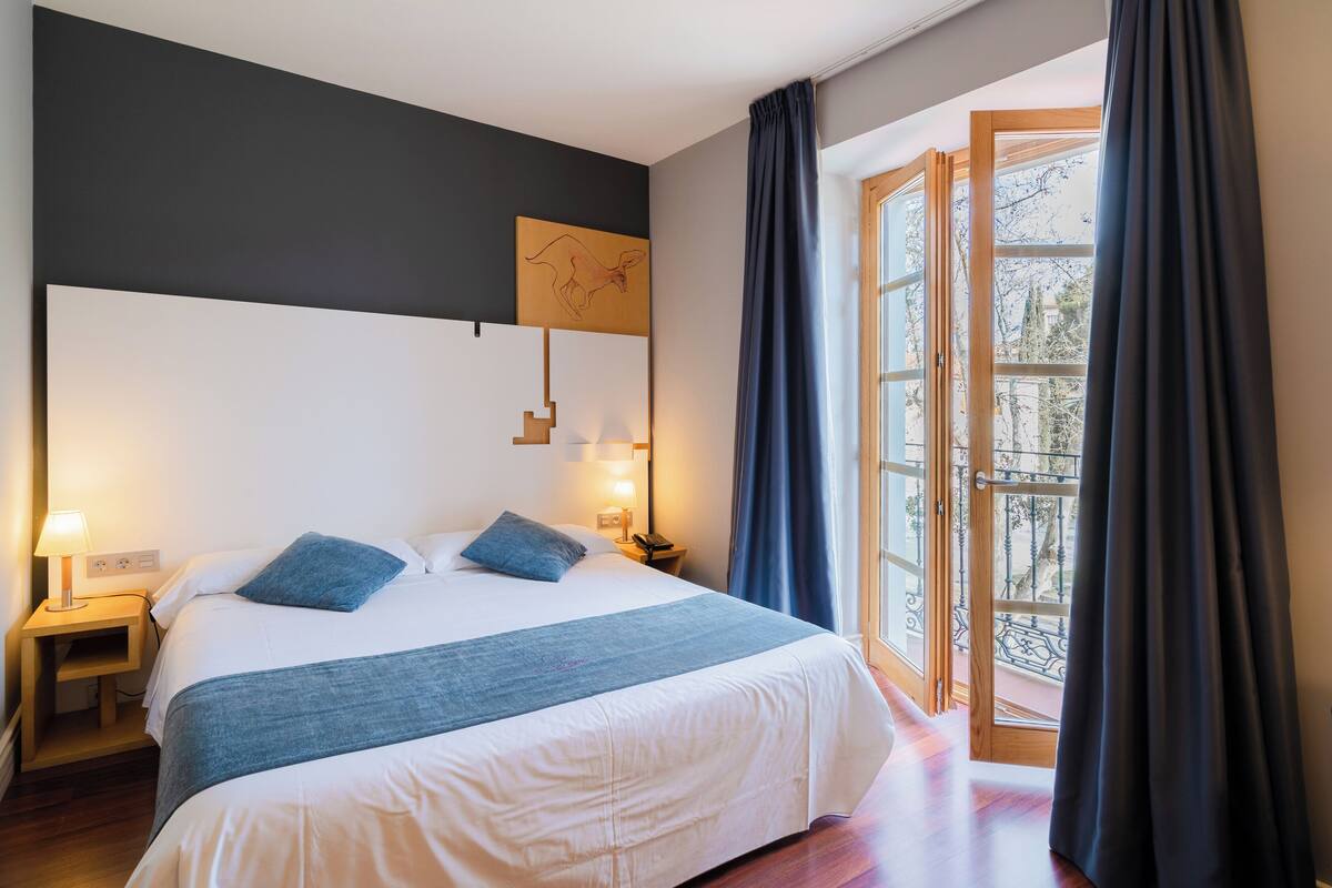 A hotel room with a large bed, a nightstand, a lamp, a map artwork on the wall, and a view of the outdoors through a glass door.