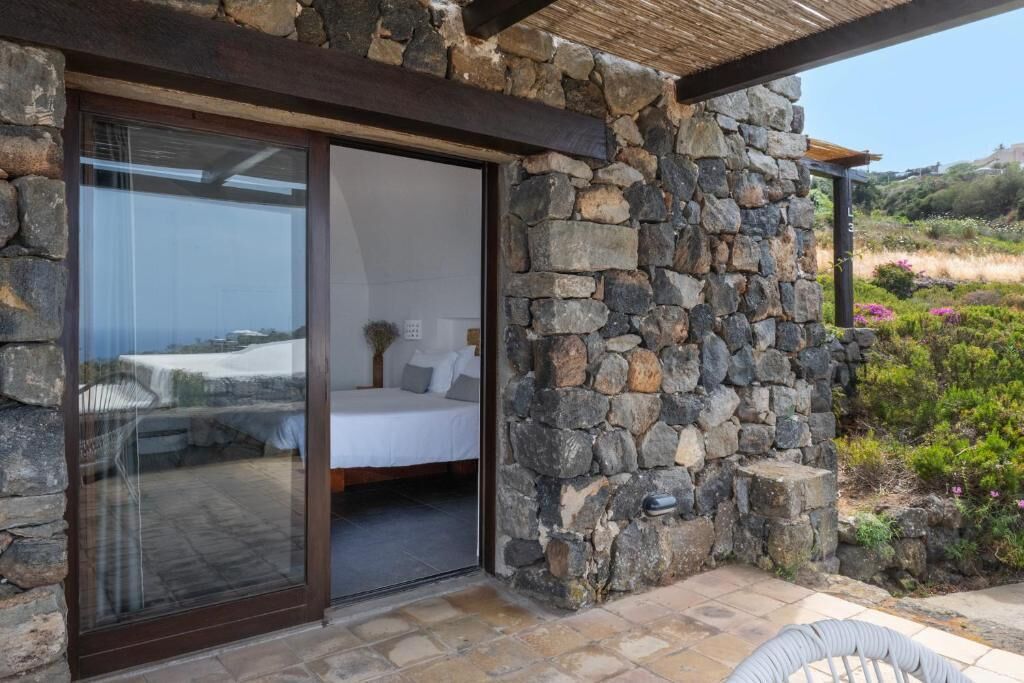 A stone exterior with a sliding glass door leading to a bedroom with a bed and a small table.