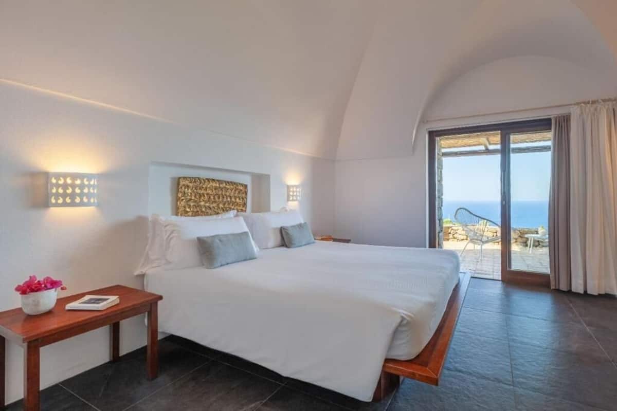 A bedroom with a large bed, a nightstand, a wall-mounted light fixture, and a view of the outdoors.