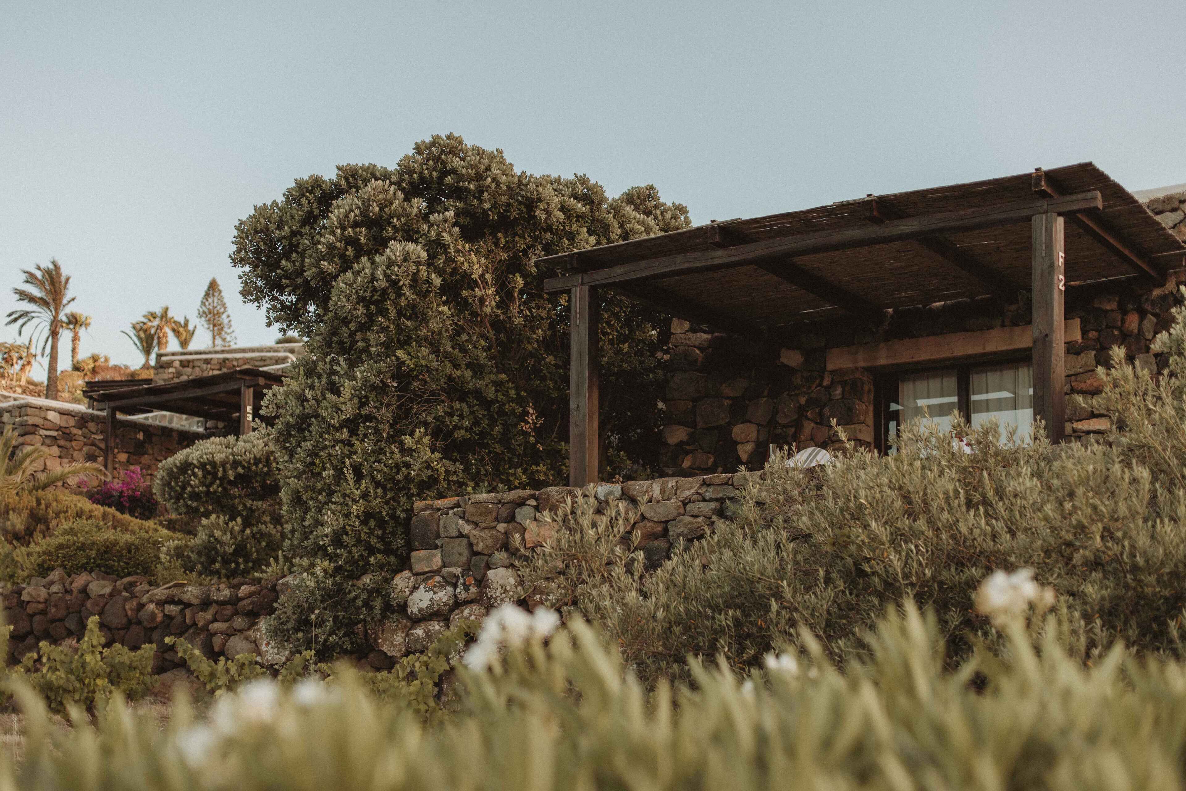 A rustic stone house with a wooden pergola, surrounded by lush greenery and palm trees.