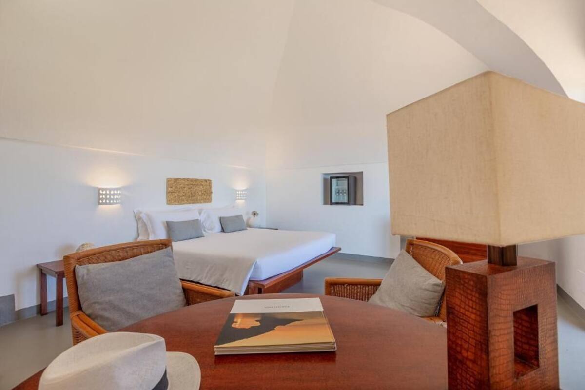 A bedroom with a bed, two bedside tables, a nightstand, a round wooden table with a magazine, and a wall-mounted lamp.
