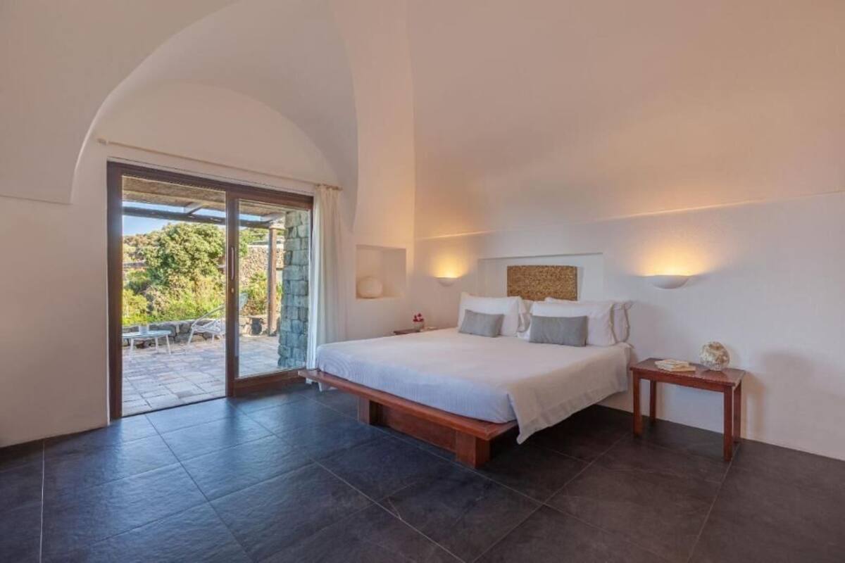 A bedroom with a large bed, a small bedside table, a wall-mounted light, and a sliding glass door leading to an outdoor area.