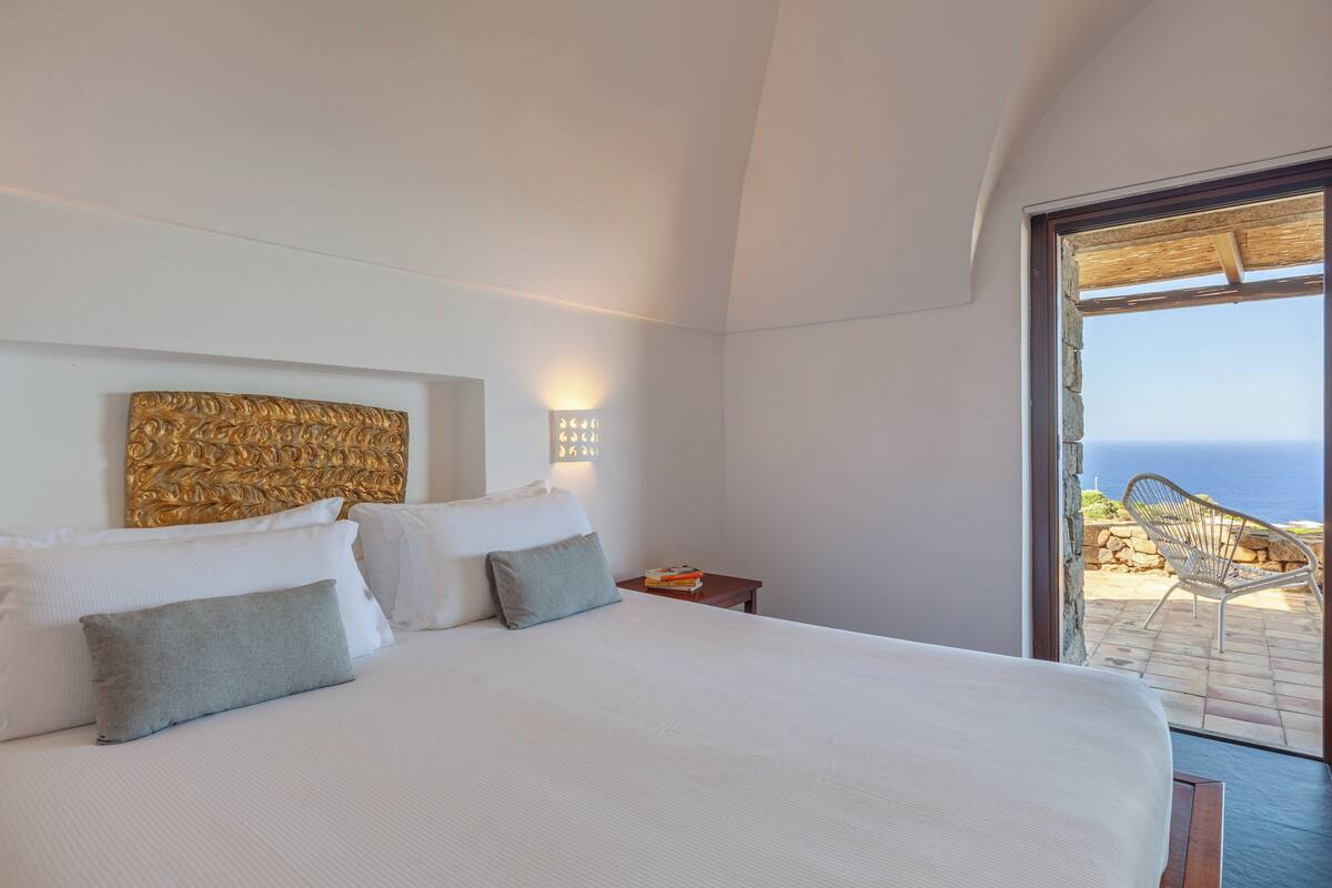 A bedroom with a large bed, a headboard, bedside lamps, and a view of the sea through an open door.