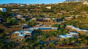 A coastal village with stone buildings, some with blue roofs, and a prominent swimming pool area.