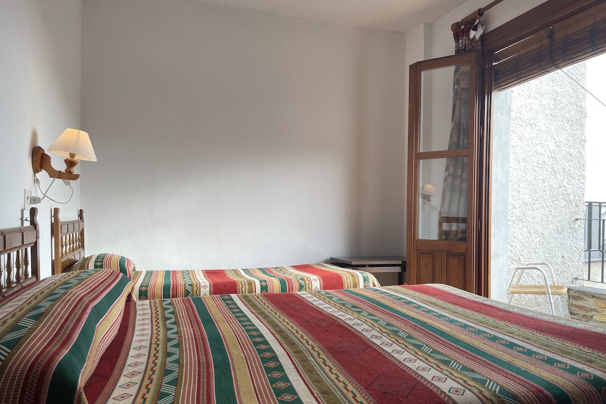A room with two beds, a wooden headboard, a wall-mounted lamp, and a large window with a view of a building.